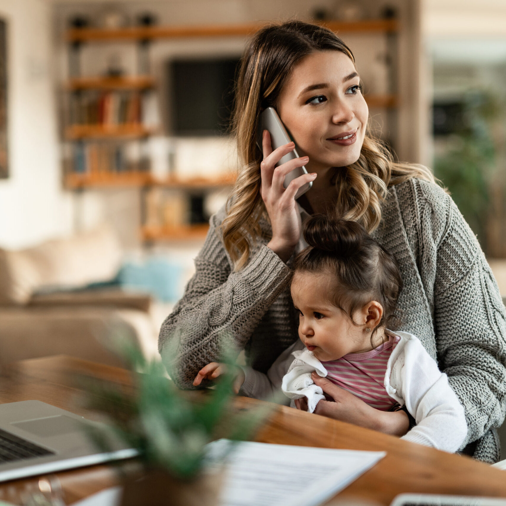 Smiling mother holding small daughter in her lap while talking on the phone at home.